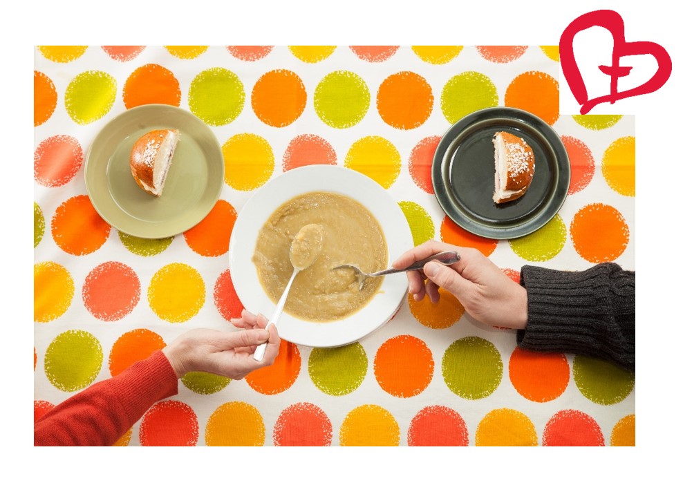 Värikkäällä pöytäliinalla hernekeittolautanen, josta kaksi ihmistä syö yhdessä. Kaksi lautasta, jossa puolikas laskiaispulla kummassakin. Yhteisvastuusydän oikeassa kulmassa.