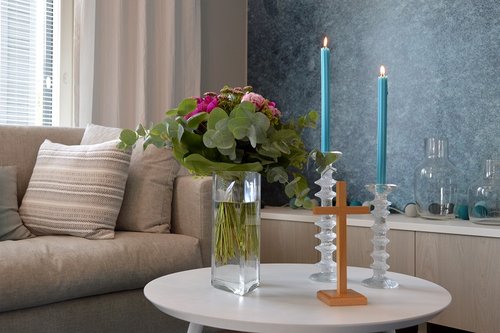 Vaalea sohva ja vaaleat verhot. Sohvapöytä, jonka päällä on kukkamaljakko, puuristi ja kaksi festivo kynttiläjalkaa sinisillä pitkillä kynttilöillä.